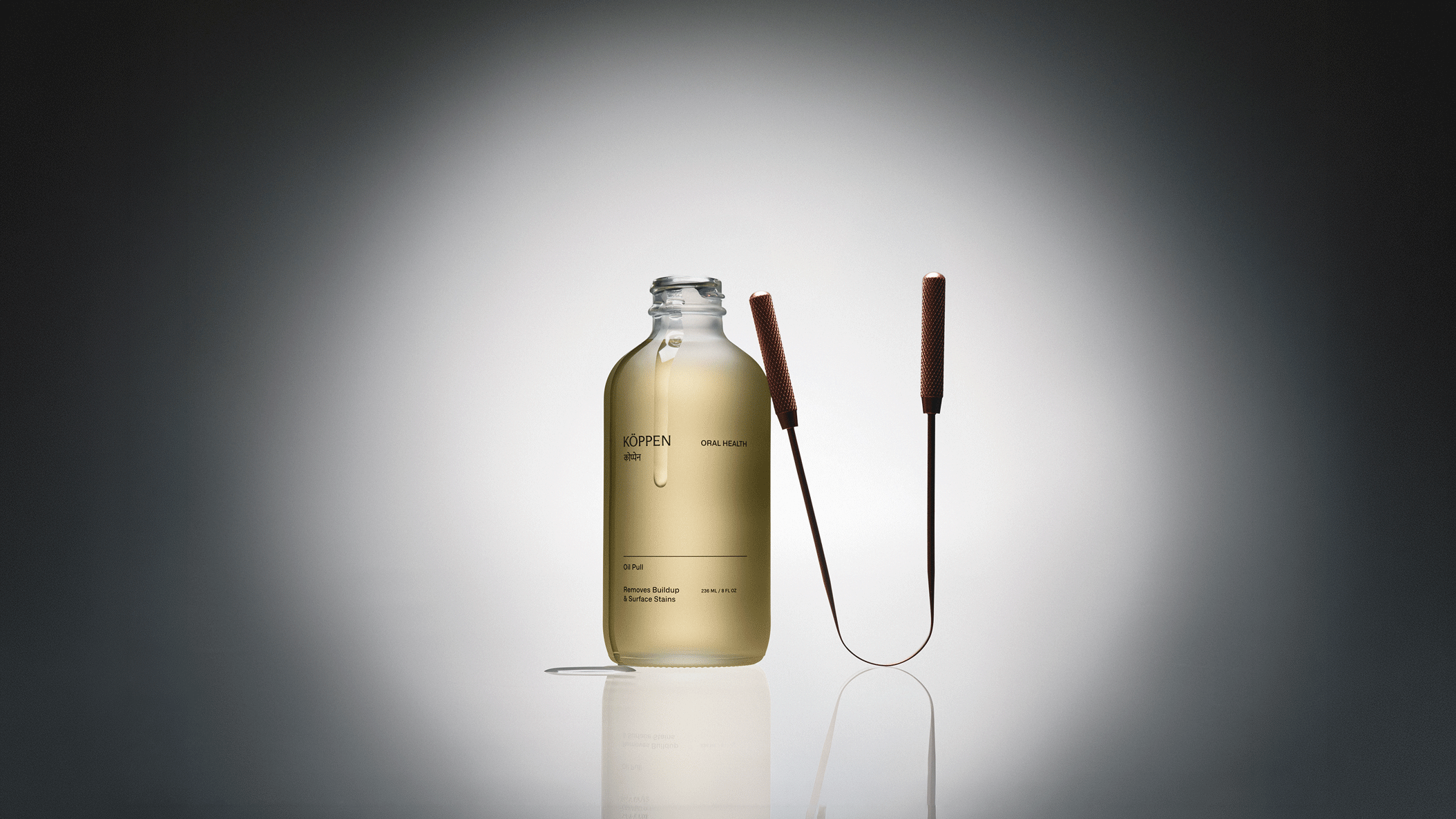 Clear frosted bottle with yellow liquid and copper tongue scraper leaning against the bottle on a gray background