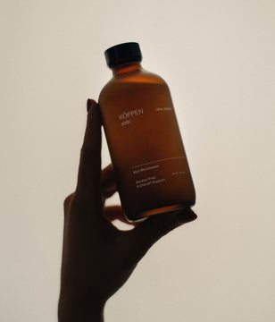 Hand holding a brown amber frosted bottle labeled 'Köppen' against a plain background